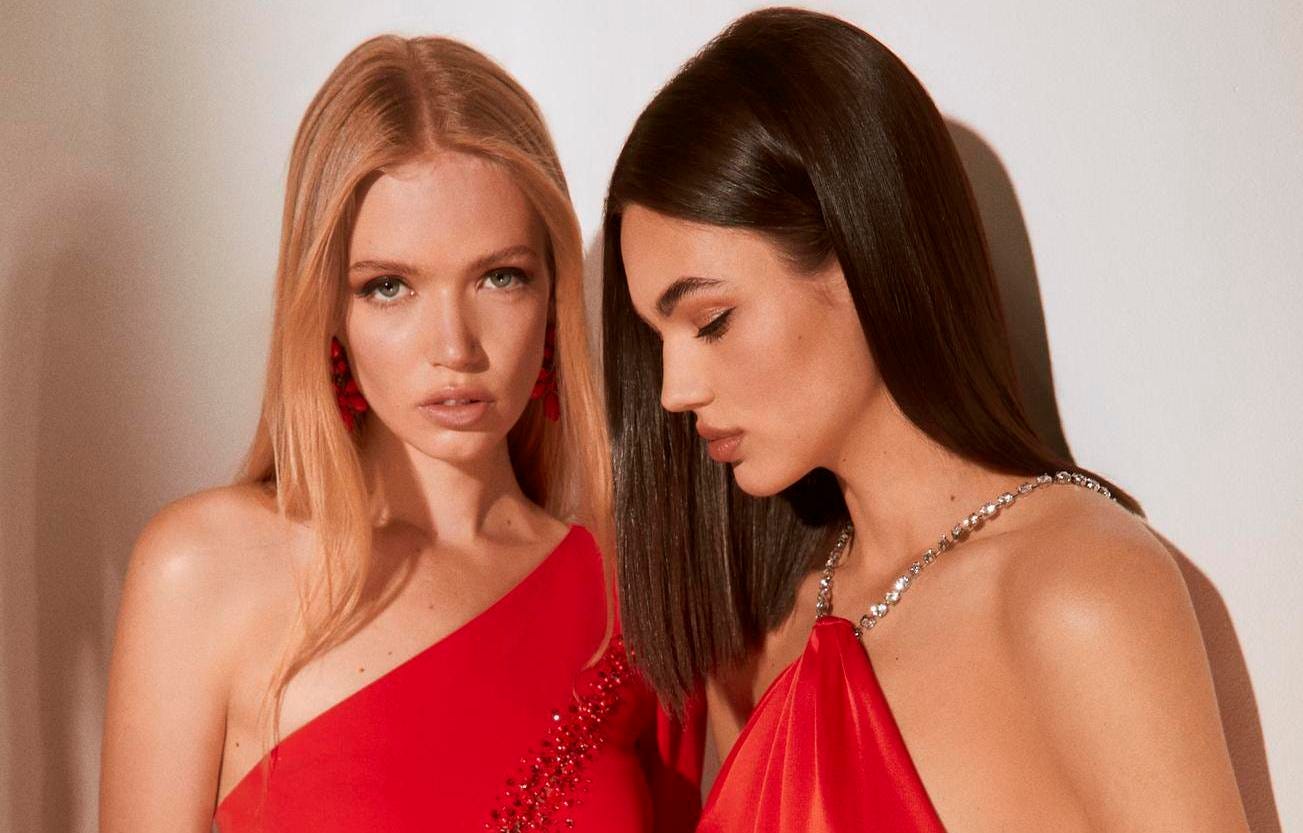 Two women wearing red bridesmaid dresses, one with a halter neck and one in a one-shouldered design.