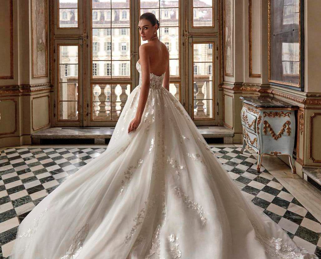 Brunette woman in a ball gown wedding dress standing in a ball room and looking back at the camera.