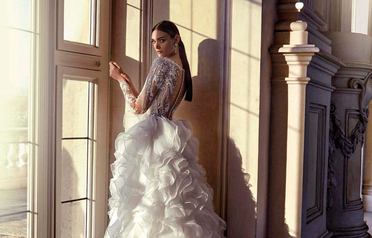 Modella di spalle che indossa un abito da sposa a maniche lunghe decorate e gonna lunga voluminosa.