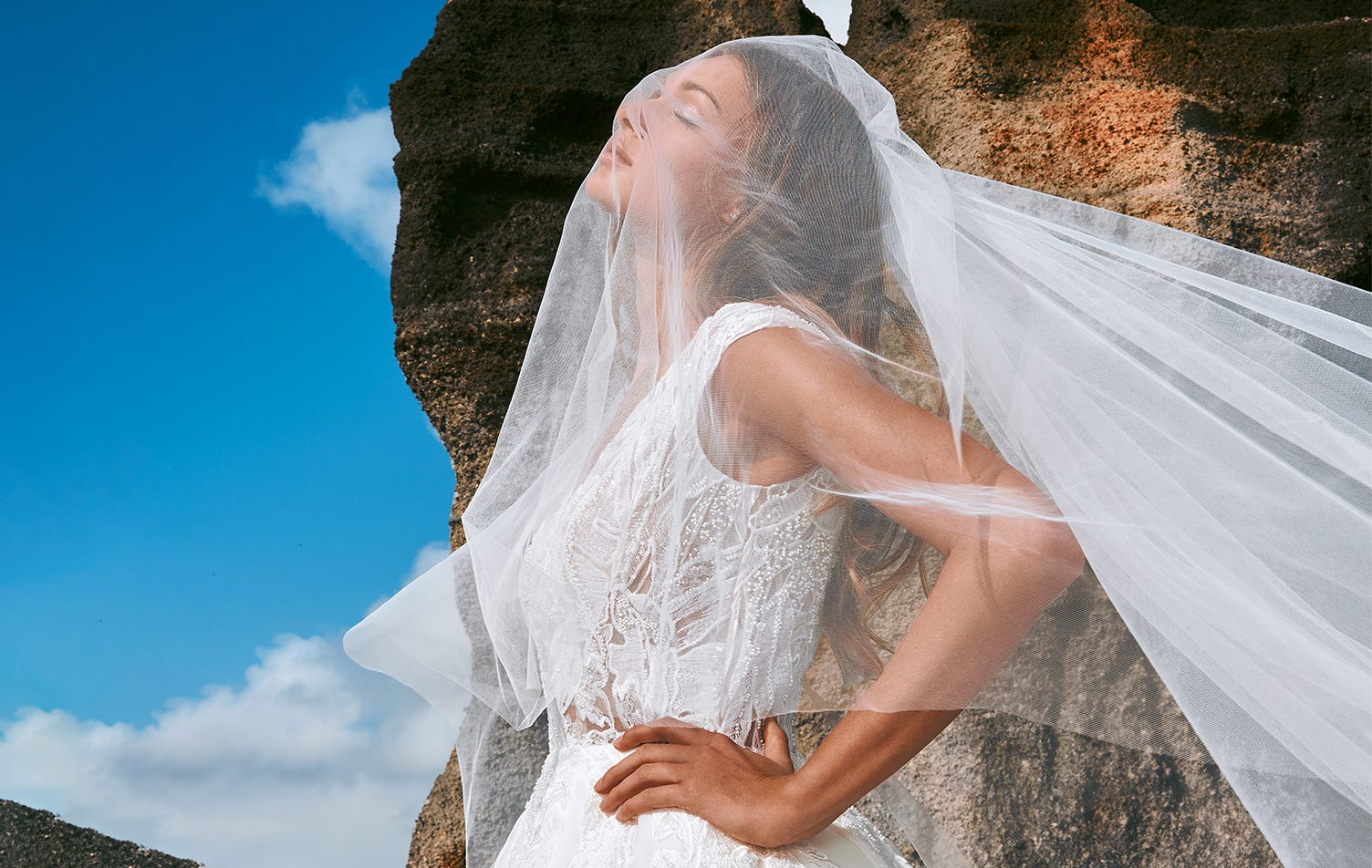 Mujer al aire libre vestida de novia con los ojos cerrando con un velo de novia cayéndole por la cara