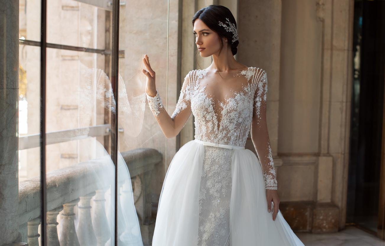 Brunette wearing a mermaid wedding gown with long sleeves and an overskirt, looking outside the window in a ballroom