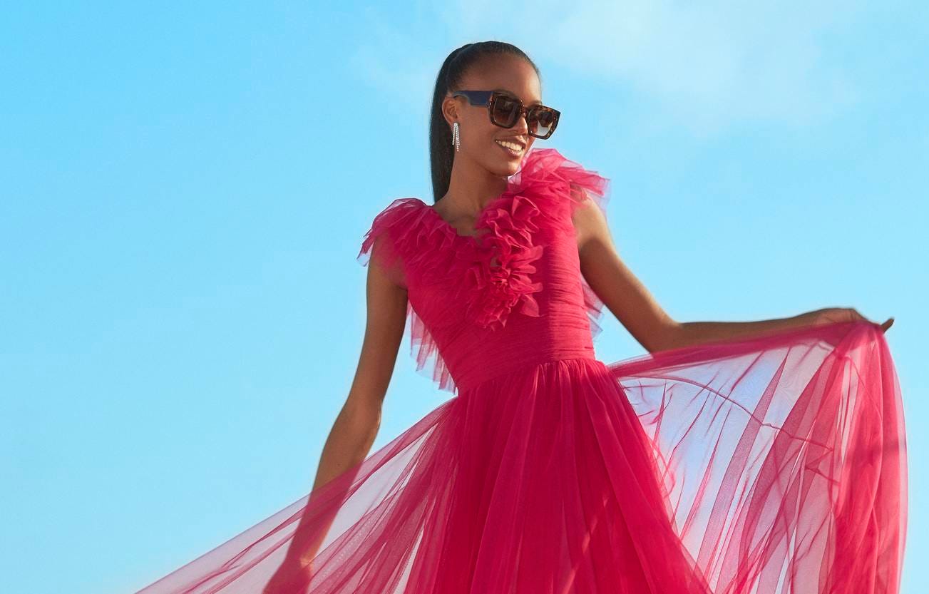 Woman wearing a pink A-line party dress with V-neck and sunglasses, standing outside and holding her gown