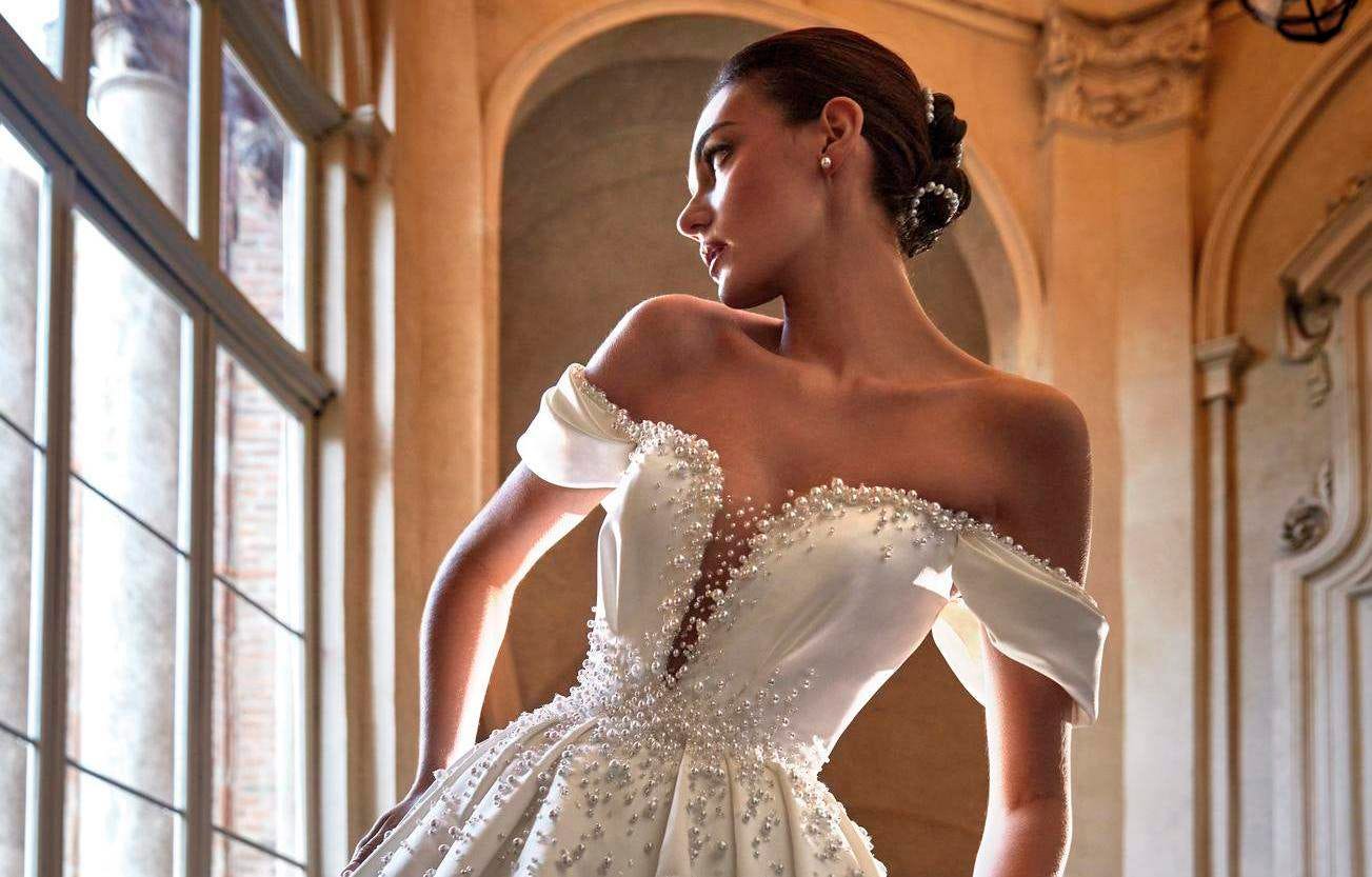 Woman in an off-the-shoulder bridal gown with intricate beading along the neckline and hair in a low bun.