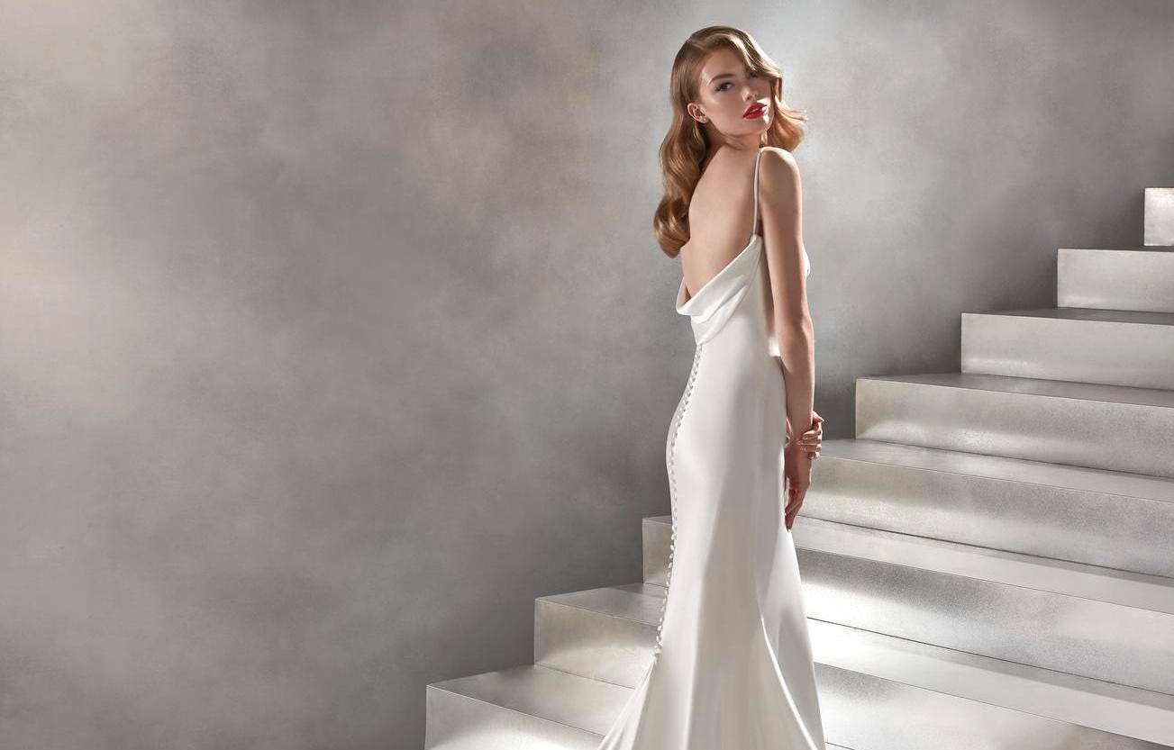 Woman wearing a satin wedding dress with wavy hair and red lipstic, standing on a white staircase