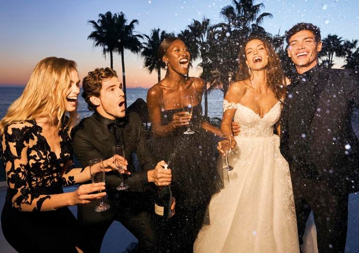 Dark-haired woman celebrating in an off-the-shoulder wedding dress with bridal party and husband dressed in black.