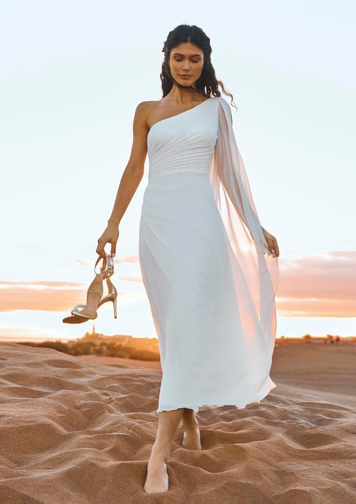 Brunette wearing a sheath bridal dress and carrying a pair of white wedding heels, walking on the beach